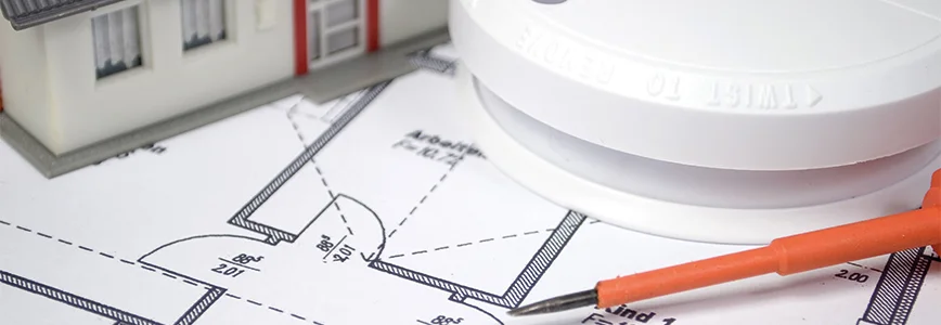 Ein Rauchmelder liegt auf einem Bauplan. Neben dem Rauchmelder liegt ein orangefarbener Schraubendreher.